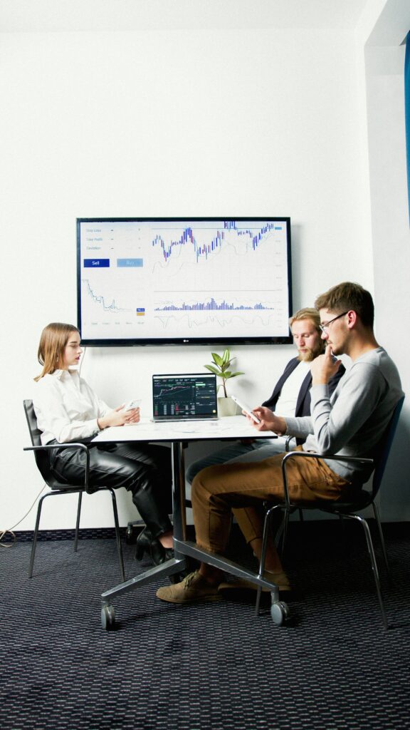 Three business professionals in a meeting room discussing financial charts displayed on a screen.